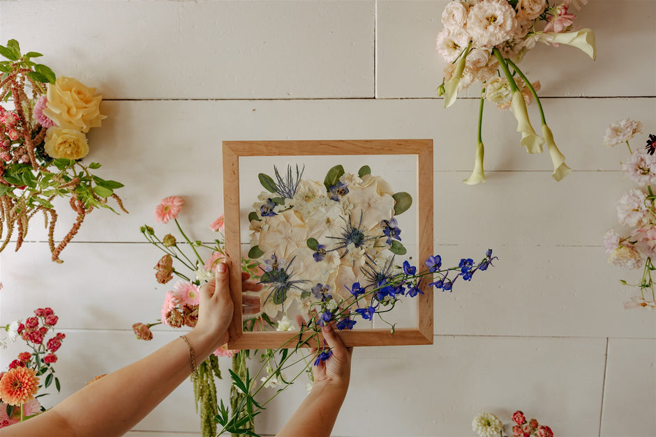 Person holding a wooden frame with a pressed floral arrangement against a white wall with hanging flowers.