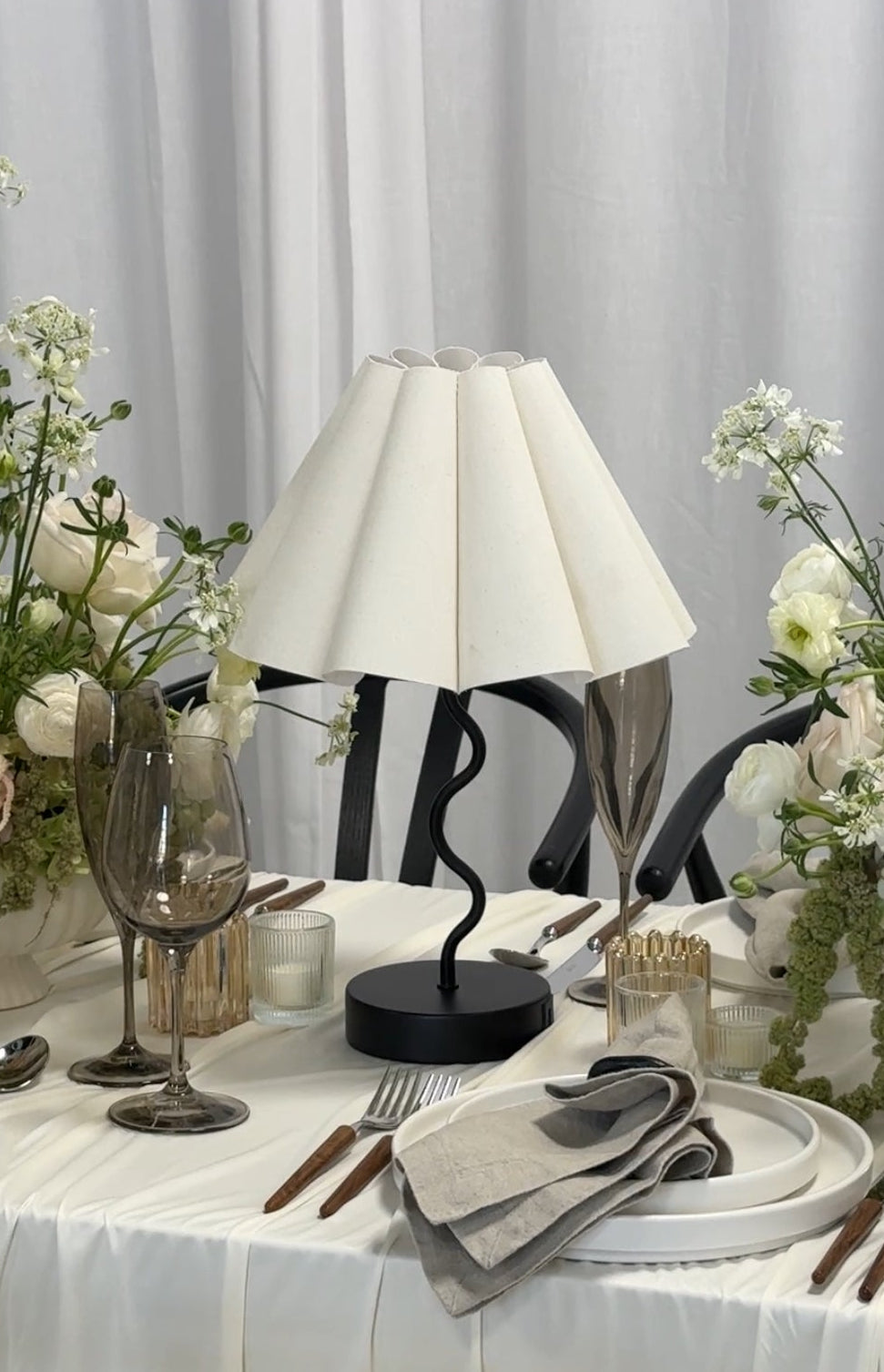 Elegant table setting with a lamp, flowers, and cutlery on a white tablecloth.