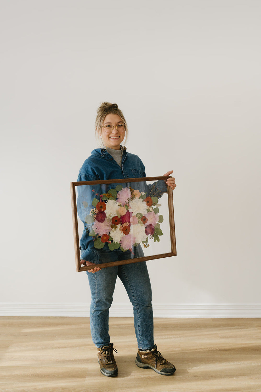 Person holding a framed floral artwork against a plain wall.