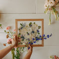 Load image into Gallery viewer, Person holding a wooden frame with a pressed floral arrangement against a white wall with hanging flowers.
