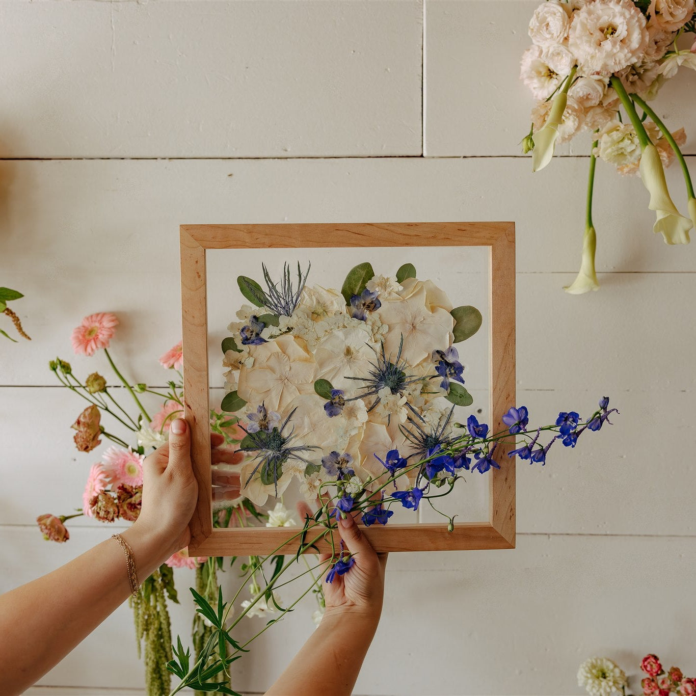 Person holding a wooden frame with a pressed floral arrangement against a white wall with hanging flowers.