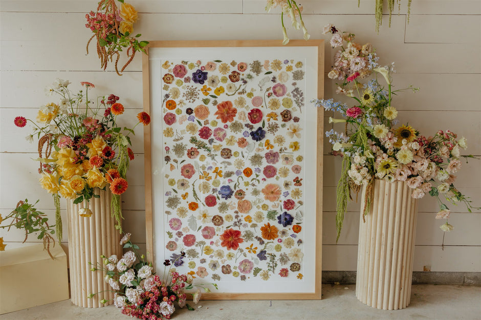 Framed pressed floral arrangement with vases of flowers on a wooden floor.